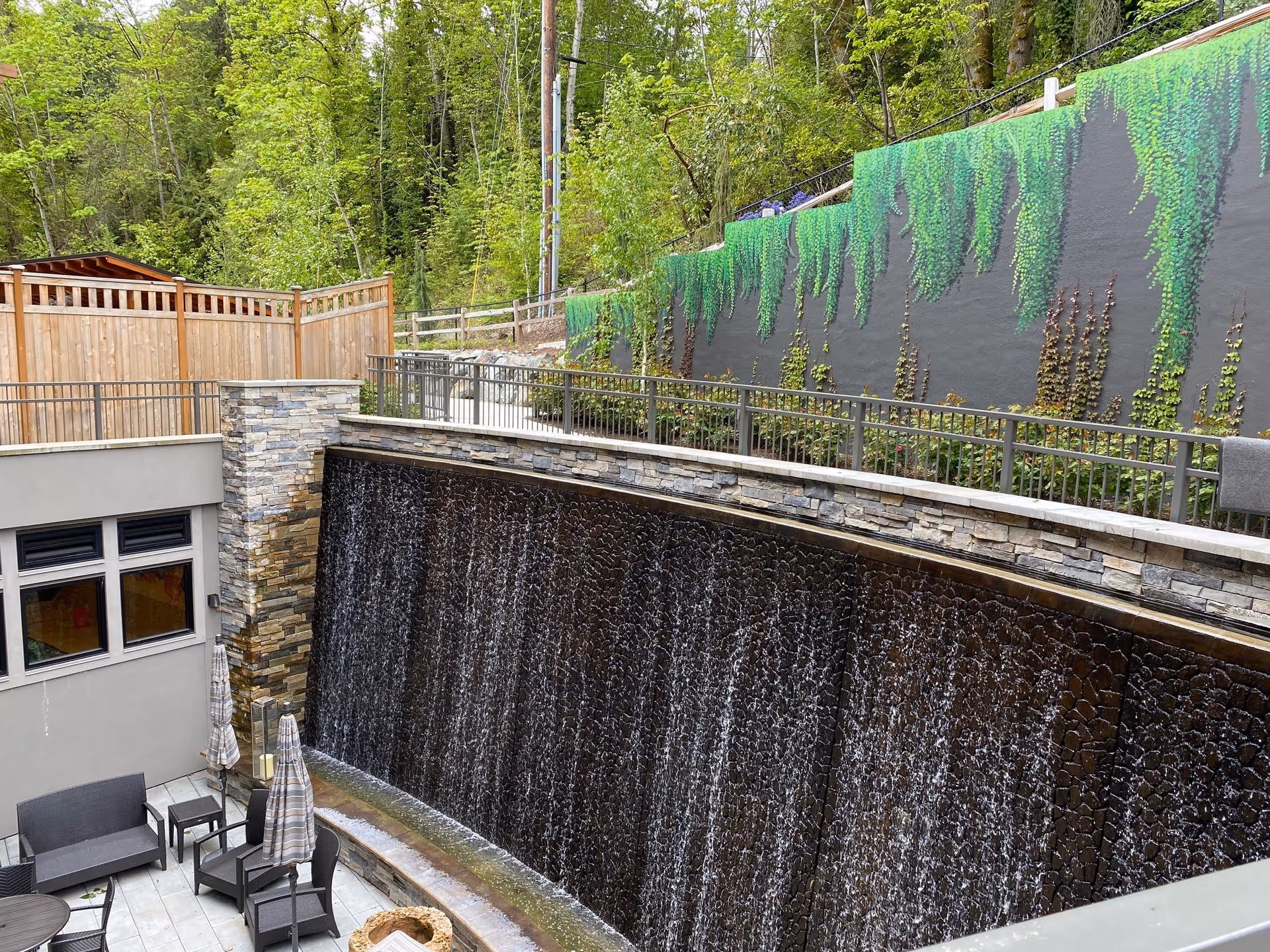 Outdoor patio area with seating including chairs and tables, adjacent to a large stone waterfall feature. The waterfall flows down a textured stone wall with greenery and a mural of hanging plants on a dark wall above. Surrounding the area are trees and a wooden fence.