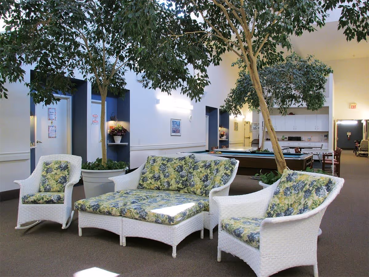 Indoor common area with white wicker furniture featuring floral cushions, large potted trees, a pool table, and a foosball table in the background. The space has white walls with blue accents and overhead lighting.