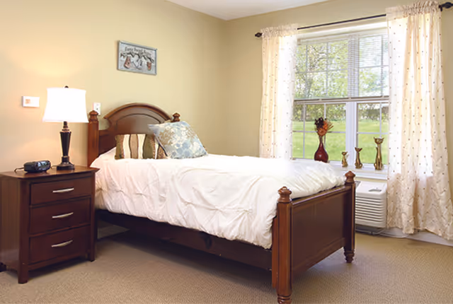 A neatly made single bed with a wooden headboard and footboard in a bedroom. The bed has white bedding and decorative pillows. Next to the bed is a wooden nightstand with a lamp and a clock. A window with light-colored curtains lets in natural light, and there are decorative items on the windowsill.