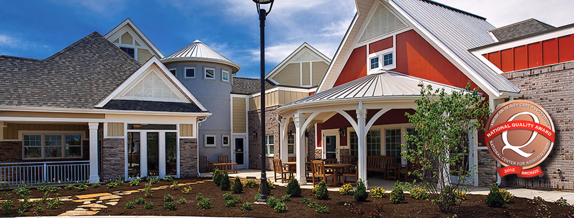 Exterior view of a senior living facility with multiple connected buildings featuring varied architectural styles, including a red building with a covered porch, a round tower-like structure, and stone accents. There is a landscaped garden with small shrubs and a stone pathway in the foreground under a partly cloudy sky.