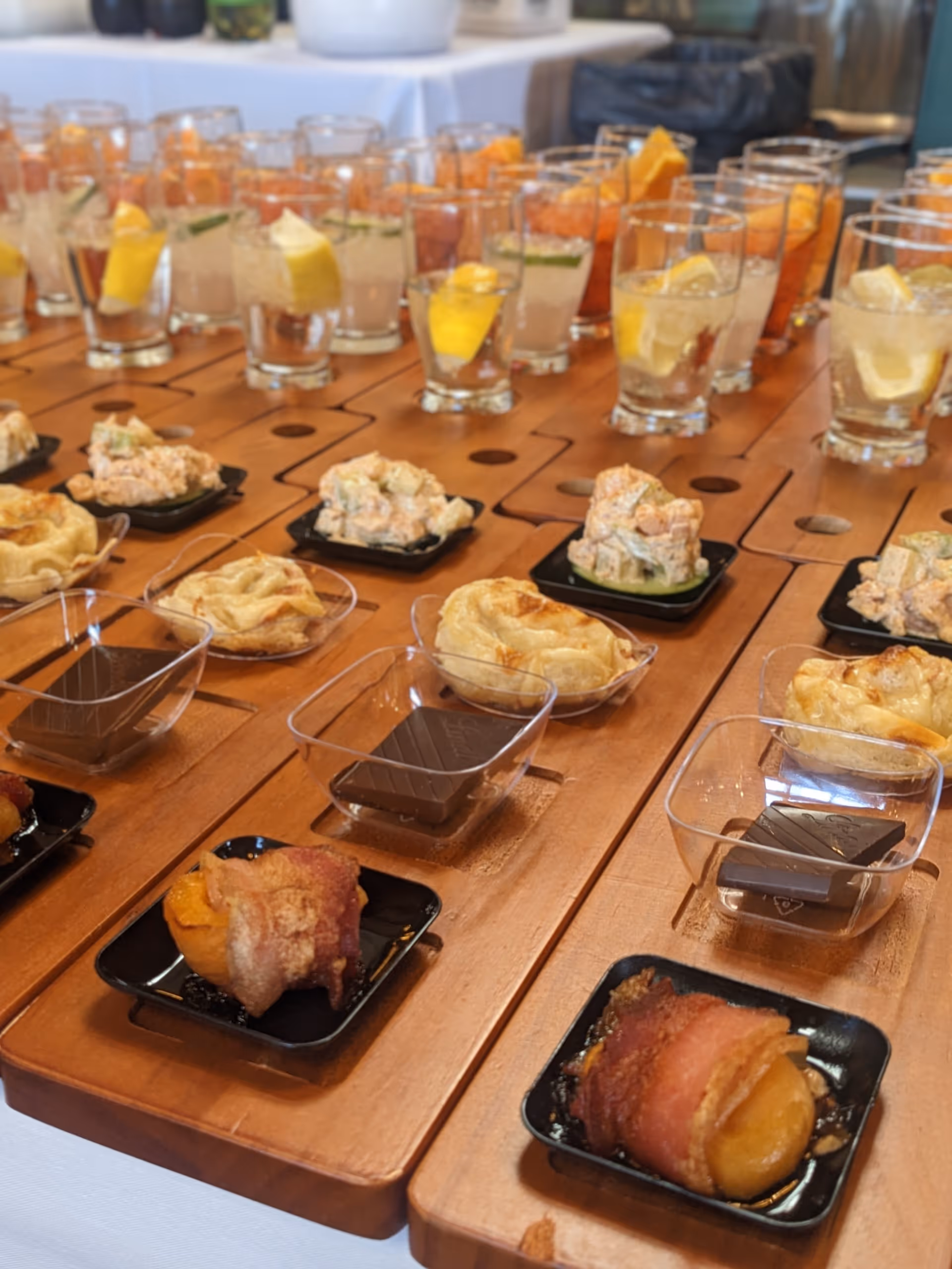 Close-up view of a wooden serving board with an assortment of appetizers and drinks. The appetizers include bacon-wrapped items, small pastry cups, salad portions, and pieces of chocolate in clear plastic containers. Behind the appetizers, there are multiple glasses filled with beverages garnished with lemon slices and herbs.