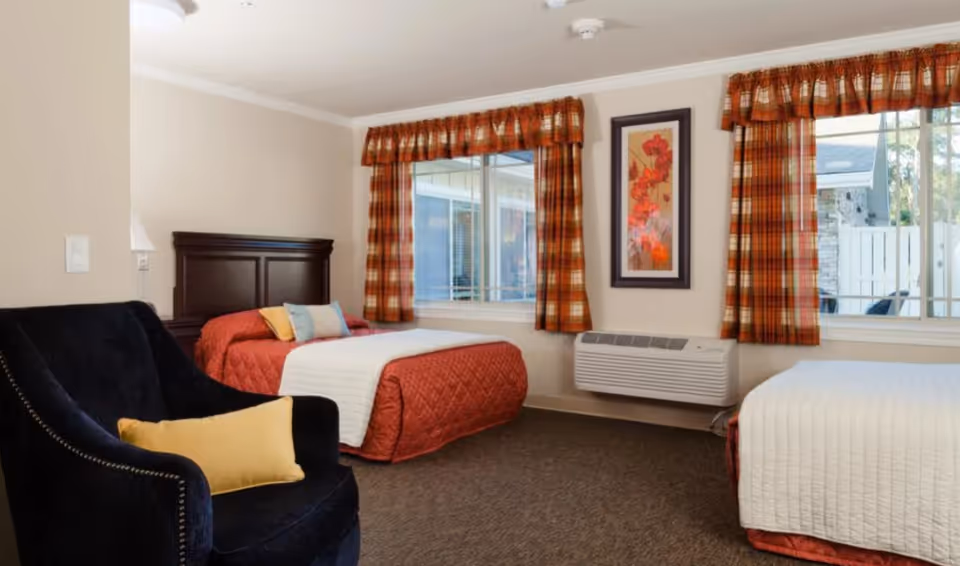 Two-bed bedroom with twin beds, a dark armchair, plaid curtains, wall art, and a window-mounted air unit.