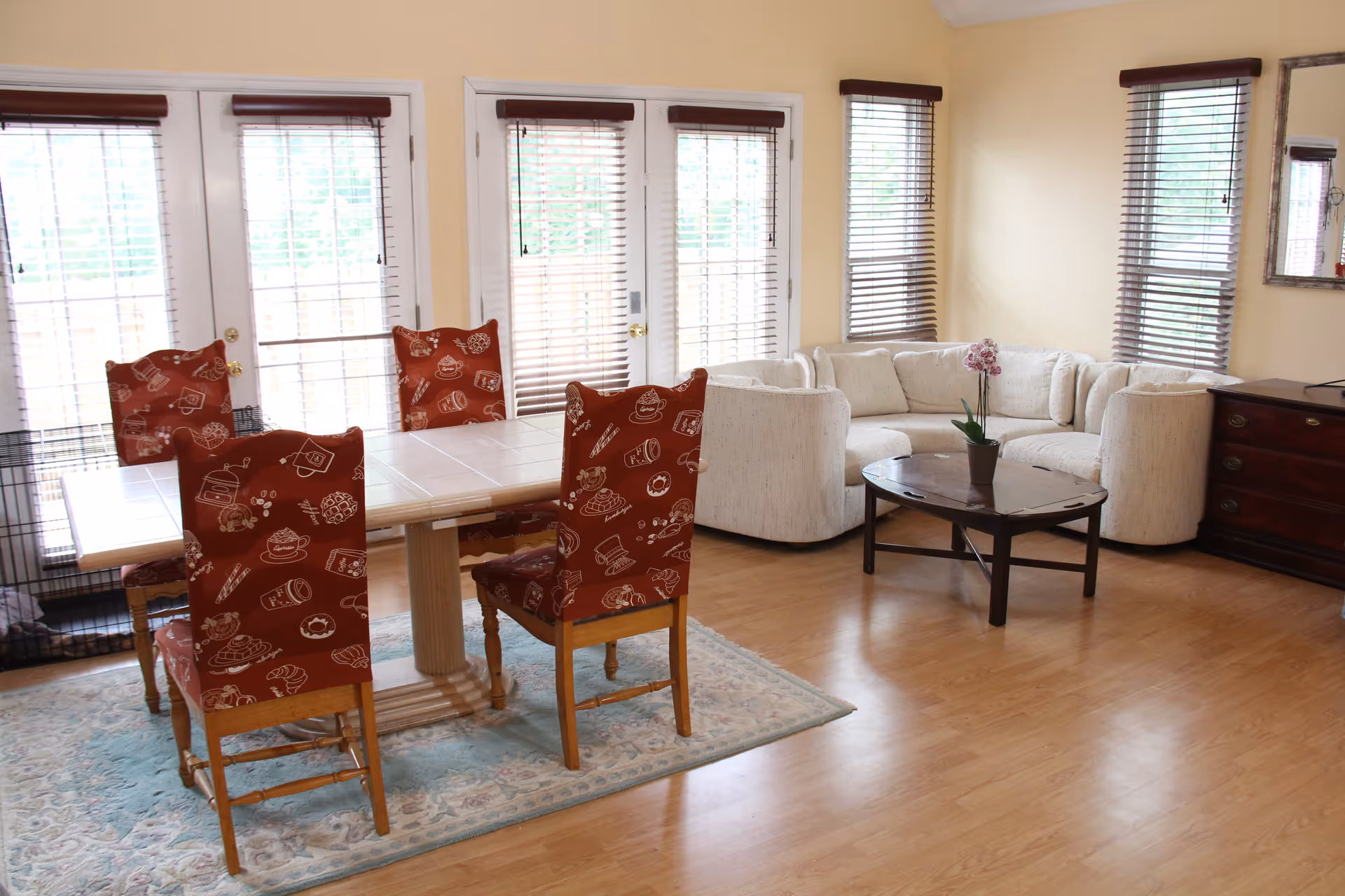 A bright living and dining area with a white sectional sofa, a wooden coffee table with a potted orchid, a dining table with four wooden chairs covered in red patterned fabric, large windows with wooden blinds, and light-colored walls and flooring.