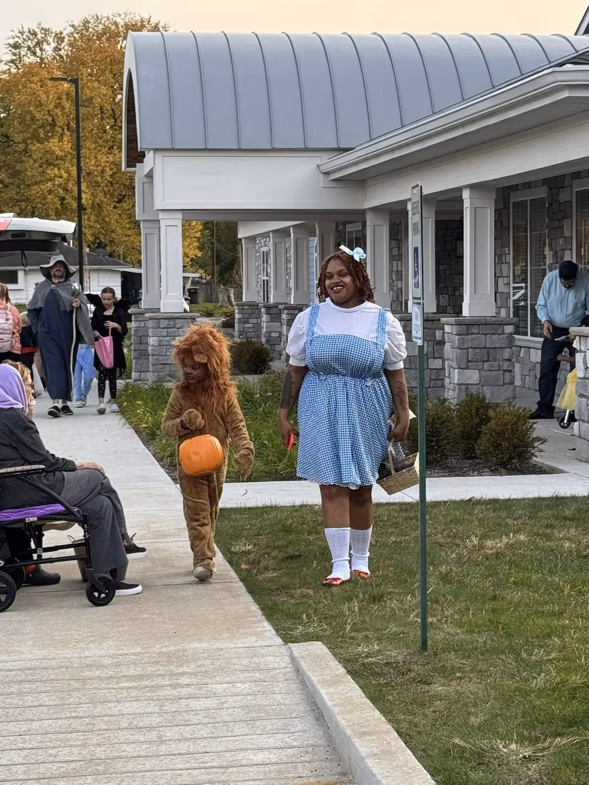 A group of people outside a building with stone and white siding. A child dressed in a lion costume holding an orange pumpkin bucket walks next to a woman dressed as Dorothy from The Wizard of Oz, wearing a blue gingham dress, white blouse, and red shoes. Other people, including an elderly person in a wheelchair and a person dressed as a wizard, are visible in the background.