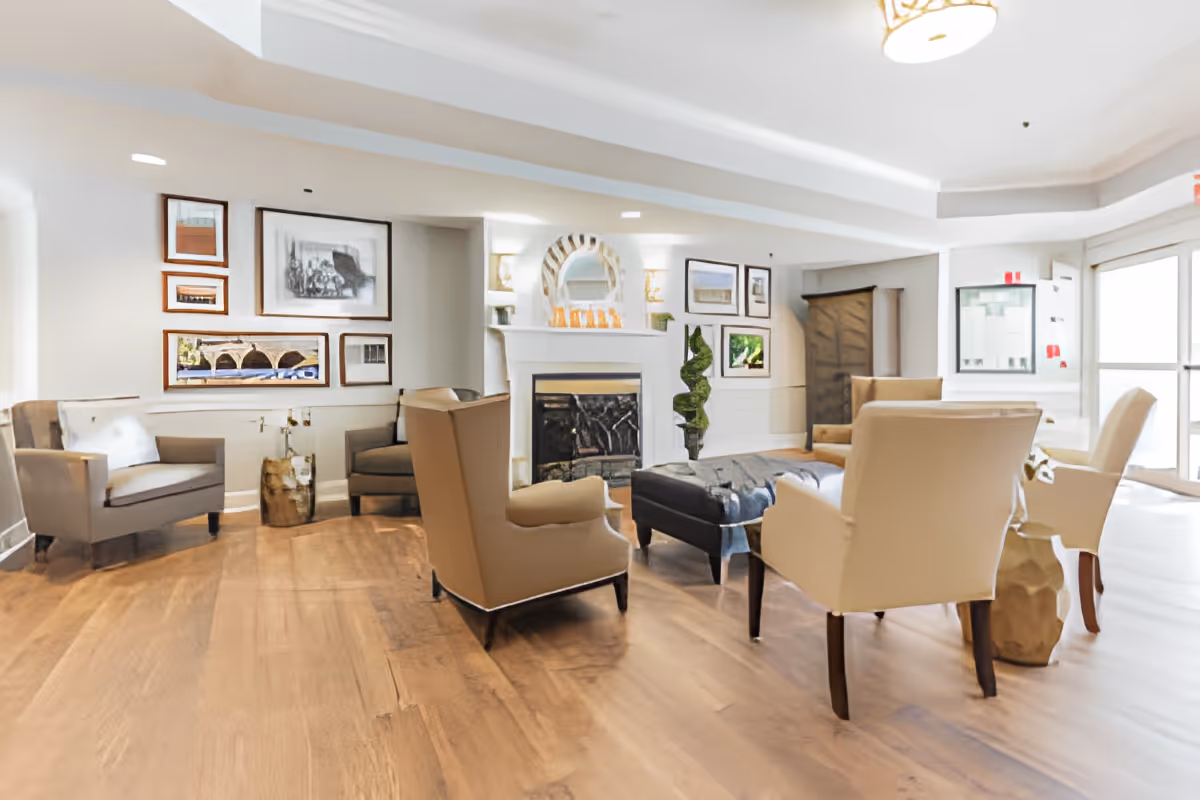 Bright communal lounge with armchairs arranged around a central ottoman and a fireplace, decorated with framed art and hardwood floors.