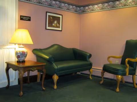 A small room with a green loveseat and a matching green armchair with wooden legs and armrests. There is a wooden side table with a decorative lamp on it, and a framed picture hanging on the wall above the loveseat. The walls are painted light purple with a floral border near the ceiling. A small sign on the wall reads 'OCCUPATIONAL THERAPY'.
