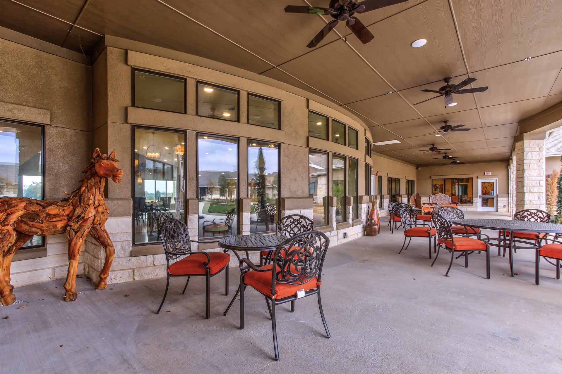 Covered outdoor patio area with multiple black metal tables and chairs with red cushions. The patio has a ceiling with several ceiling fans and large windows looking into the building. A wooden horse sculpture is positioned near the windows on the left side.