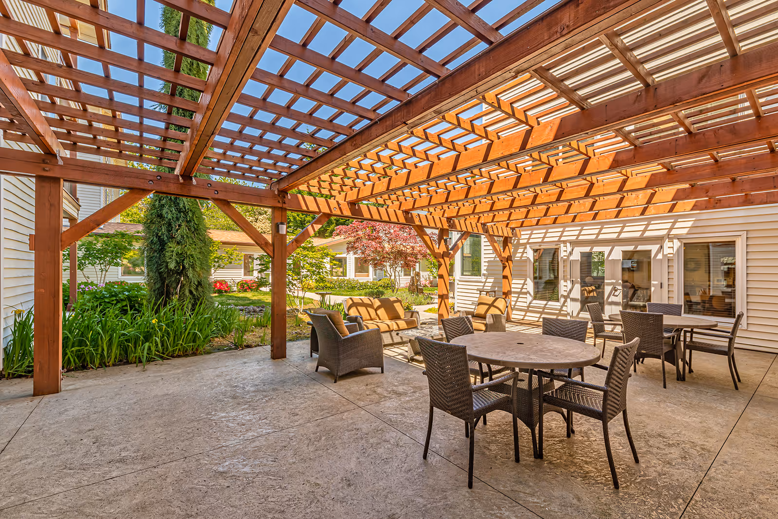 Outdoor patio area at Courtyard At Mount Tabor with a wooden pergola overhead casting shadows on the concrete floor. The space includes round tables with wicker chairs and cushioned seating, surrounded by greenery and trees with a building visible in the background.