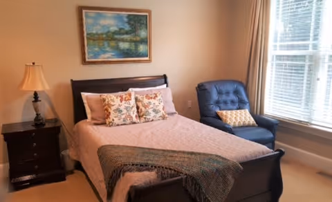 A cozy bedroom with a wooden bed frame, a pink bedspread, and two floral pillows. There is a dark wooden nightstand with a lamp on the left side of the bed. A blue armchair with a patterned cushion is placed near a large window with blinds. A framed painting of a landscape hangs on the wall above the bed.