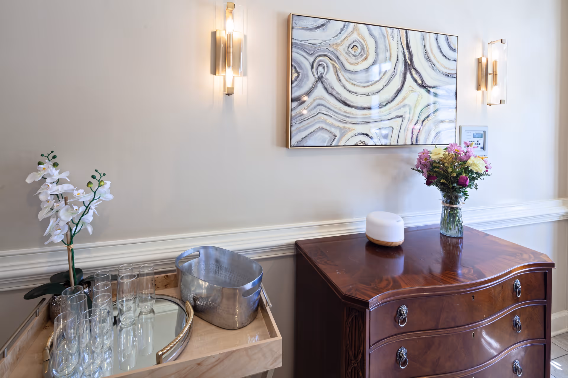A corner of a room with a wooden chest of drawers topped with a vase of colorful flowers and a small white decorative object. Above the chest hangs a framed abstract artwork with swirling patterns. On the left side, there is a wooden tray holding several empty glasses, a metal ice bucket, and a white orchid plant. Two modern wall sconces with warm lighting flank the artwork on a light-colored wall.