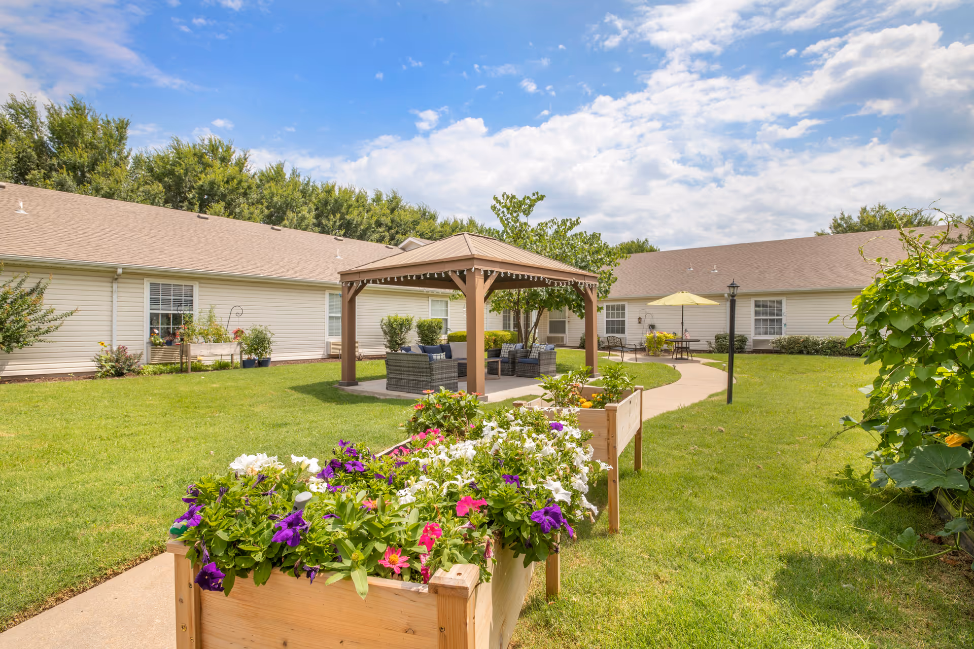 Outdoor garden area at Brookdale Broken Arrow featuring raised wooden flower beds with colorful flowers, a paved walkway, a gazebo with outdoor seating, and a building with beige siding and multiple windows under a partly cloudy blue sky.