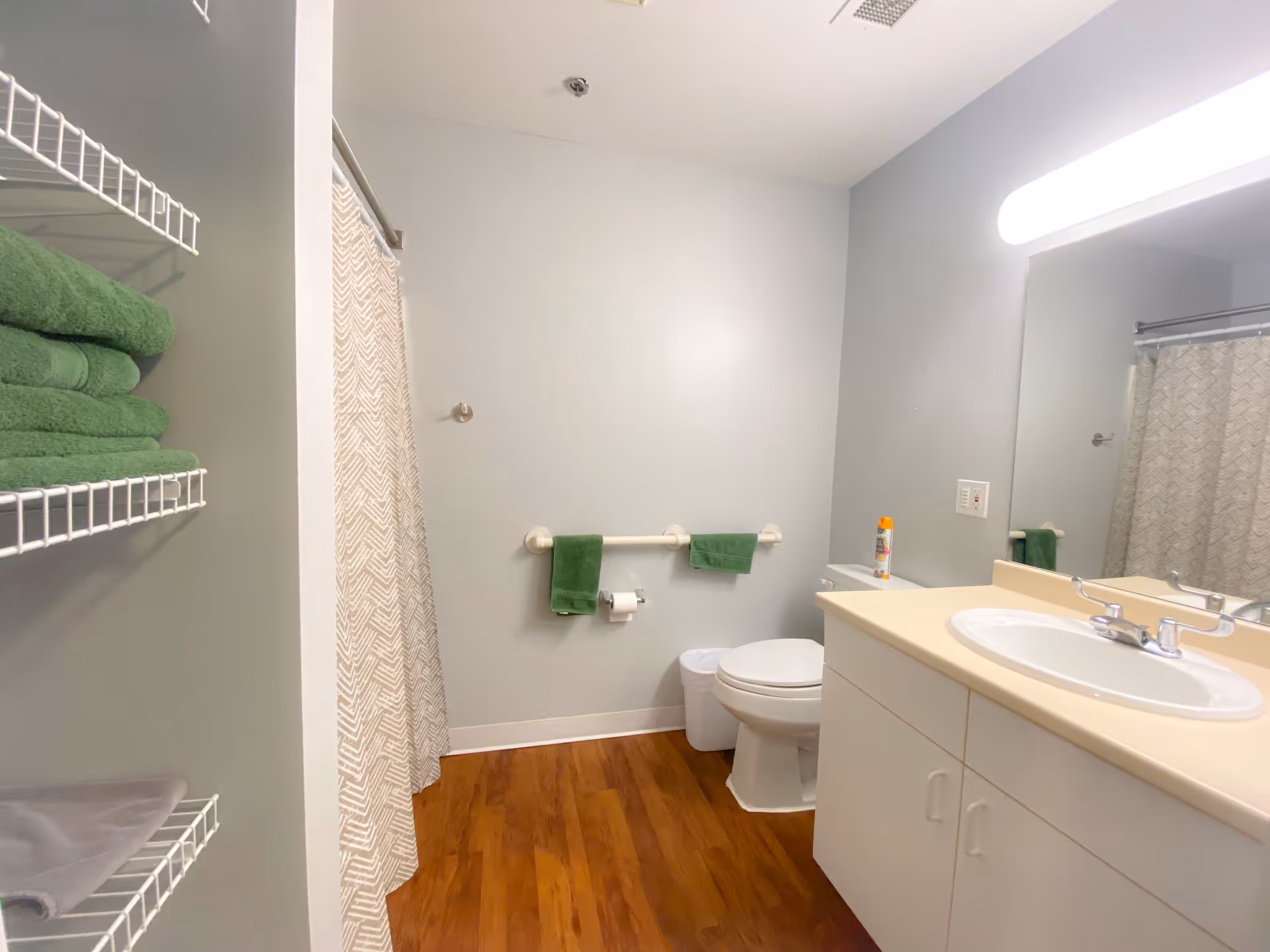 A clean bathroom with light gray walls and wood-look flooring. There is a white toilet with a white trash bin beside it, a white sink with a beige countertop and cabinet below, and a large mirror above the sink. A shower with a patterned curtain is visible on the left side. Green towels are neatly folded on white wire shelves and hanging on a grab bar next to the toilet. A can of spray is placed on the toilet tank.