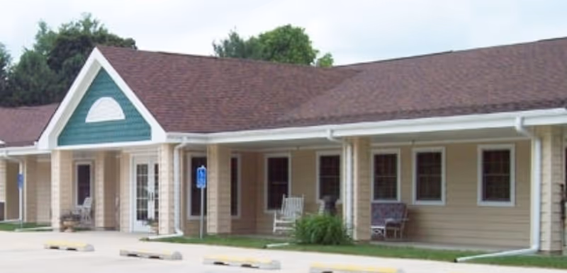 Exterior view of a single-story assisted living facility building with beige siding, a brown shingled roof, several windows, and a covered porch area with rocking chairs. There are handicap parking signs and parking spaces in front of the building, surrounded by some greenery and trees in the background.