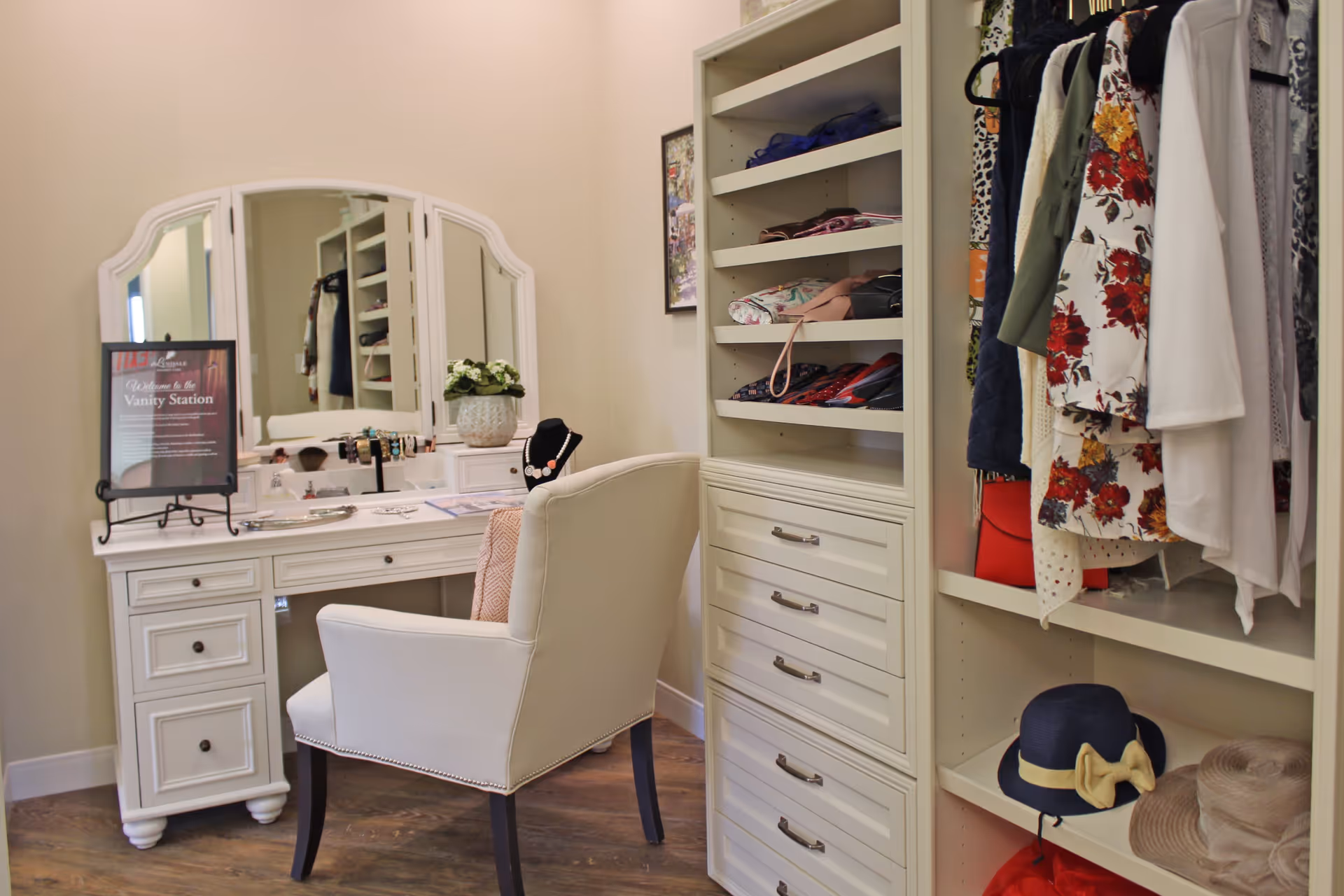A cozy vanity station with a white desk and a cushioned chair in front of a three-panel mirror. The desk holds various jewelry items, a small potted plant, and a framed sign. To the right, there is a built-in shelving unit with folded clothes, handbags, hanging garments, and hats on display.