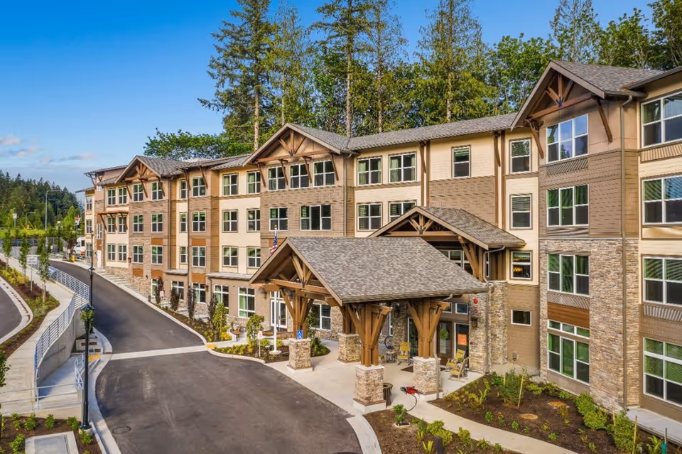 Exterior view of a multi-story senior living facility building with a covered entrance, surrounded by landscaped greenery and trees under a clear blue sky.
