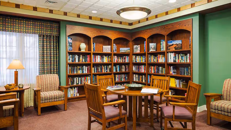 A cozy library room in a senior living facility featuring wooden bookshelves filled with books and decorative items. In the center, there is a round wooden table surrounded by four wooden chairs with cushioned seats. Two upholstered armchairs are positioned near a window with patterned curtains, next to a small wooden side table with a lamp and a clock. The walls are painted green and the ceiling has recessed lighting and a central light fixture.