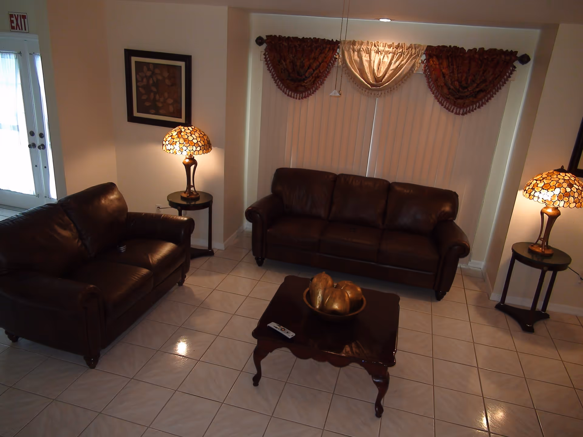 A cozy living room with two dark brown leather sofas facing each other, a wooden coffee table with decorative items, two side tables each with a lamp featuring a stained glass shade, a framed artwork on the wall, and a window with vertical blinds and decorative valances.