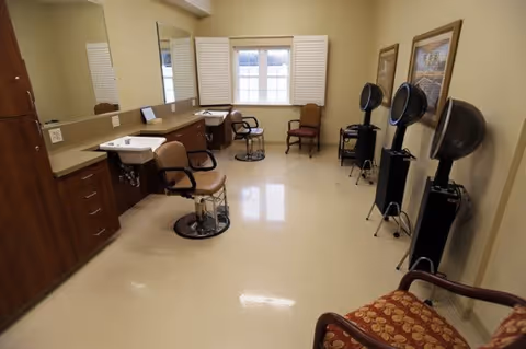 Interior view of a salon area in a senior living facility with salon chairs, hair dryers, mirrors, and a window with white shutters. The room has beige walls and a polished floor, with framed pictures on the wall and additional chairs for seating.