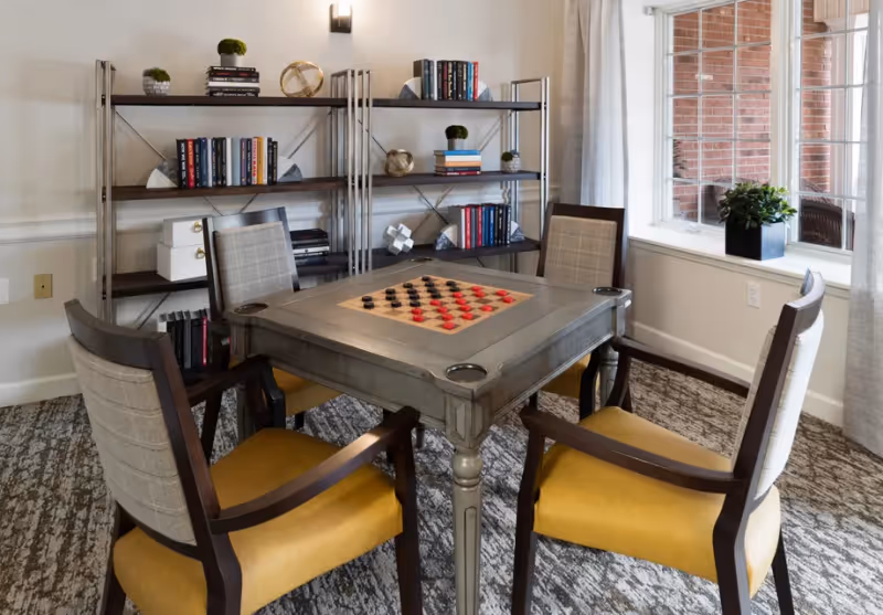 A cozy game table with a checkers board surrounded by four upholstered chairs in a lounge area with bookshelves and a window.
