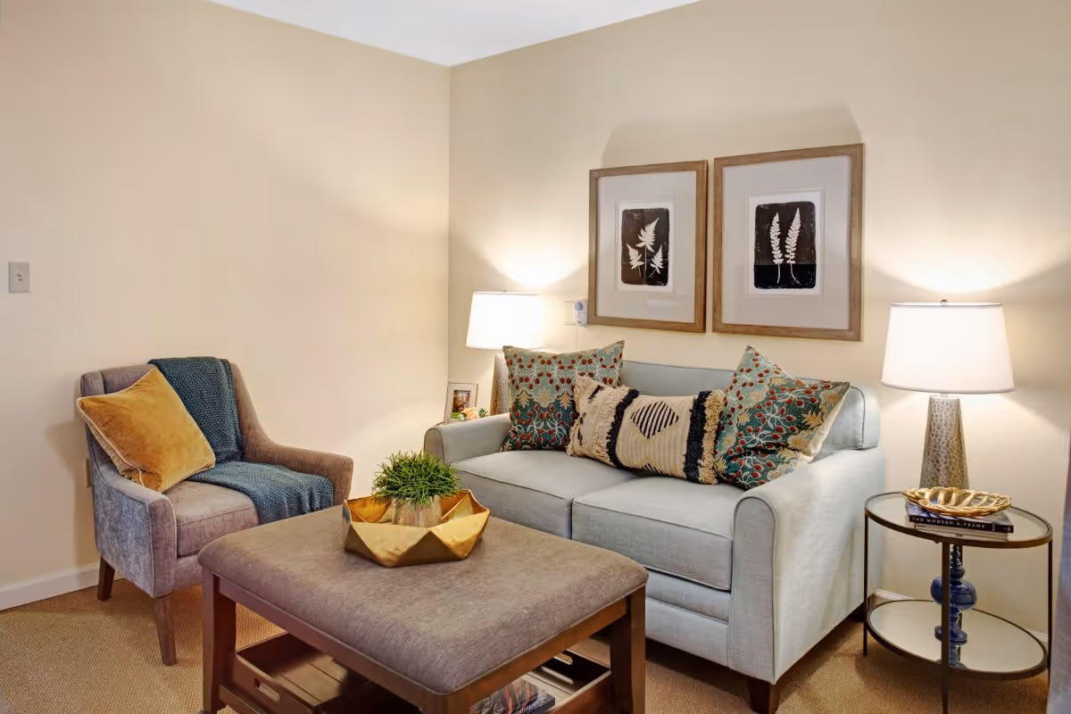 A cozy living room with a light blue sofa adorned with patterned cushions, a gray armchair with a mustard yellow pillow and a teal throw, a large ottoman coffee table with a decorative gold bowl and green plant, two framed botanical prints on the wall, and two table lamps providing warm lighting.