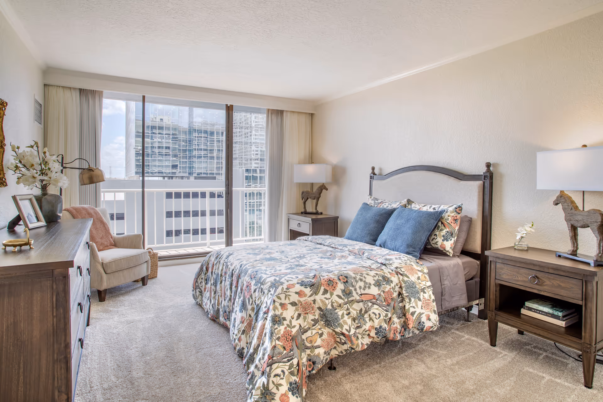 A bright bedroom with a large window showing a cityscape view. The room features a bed with floral bedding and blue pillows, two wooden nightstands with lamps and decorative horse figurines, a wooden dresser with a vase of flowers and framed photo, and a beige armchair with a pink throw blanket.