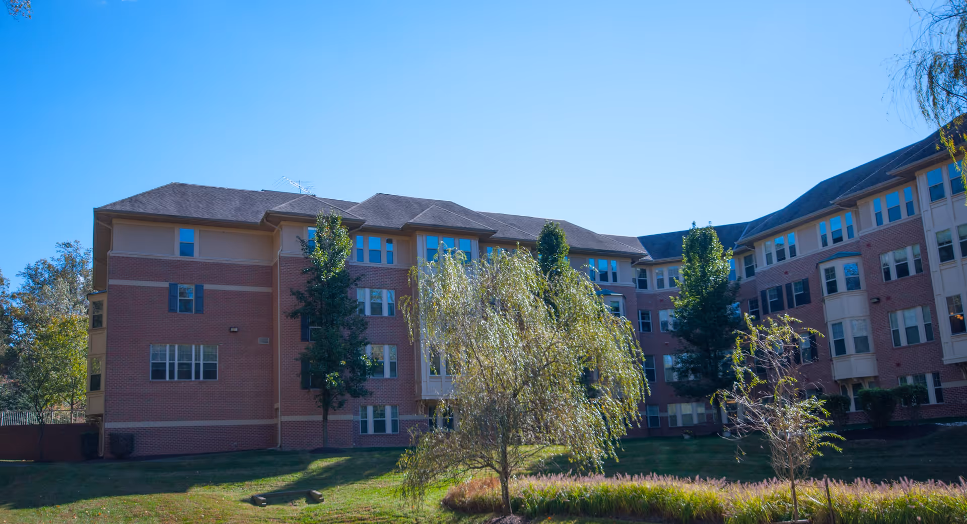 Exterior view of a multi-story brick senior living facility building with numerous windows, surrounded by green grass, trees, and landscaping under a clear blue sky.