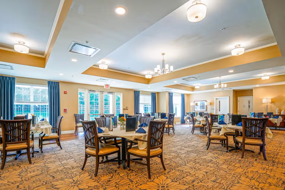 A spacious dining room with multiple tables set with white tablecloths, blue napkins, and menus. The room has large windows with blue curtains, beige walls, and a patterned carpet. Ceiling lights and chandeliers illuminate the area.