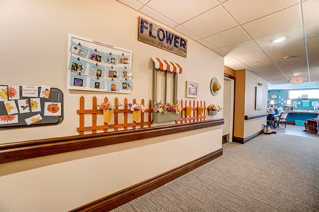 A hallway in a senior living facility decorated with a flower market theme on the wall, including a wooden sign that says 'FLOWER Market', small flower pots attached to a wooden fence decoration, and a matching flower activity board. At the end of the hallway, there are seniors sitting and engaging in activities near a window.
