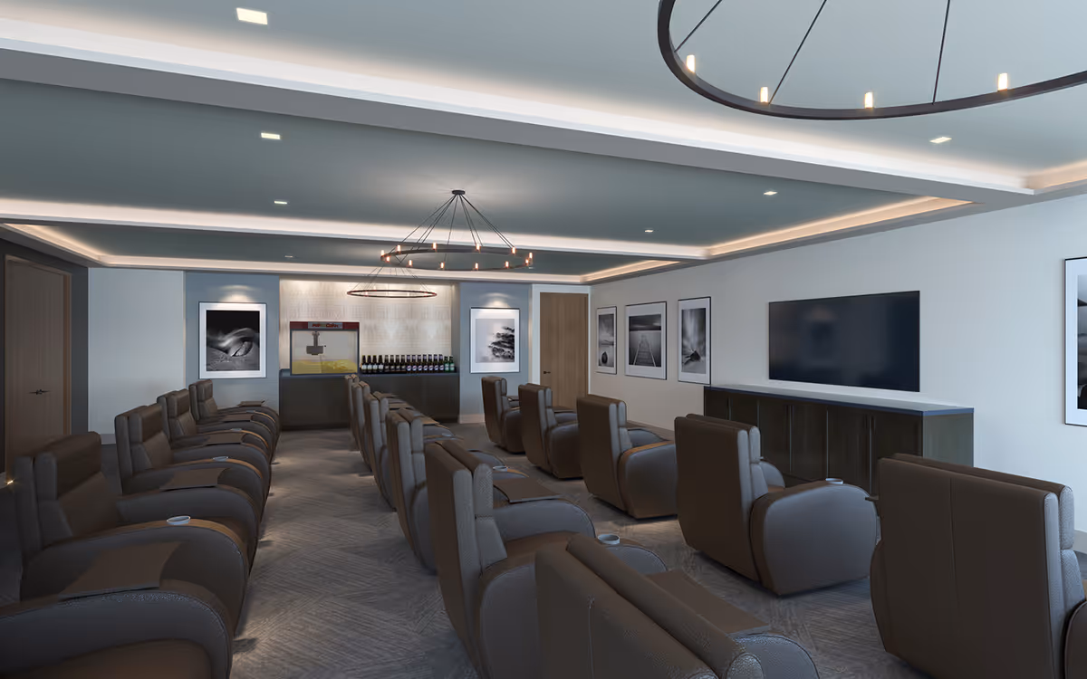 A modern lounge area with multiple rows of comfortable gray recliner chairs facing a large flat-screen TV mounted on the wall. The room features soft lighting from ceiling fixtures and a circular chandelier. There are framed black and white photographs on the walls and a popcorn machine with bottles displayed on a counter at the back of the room.