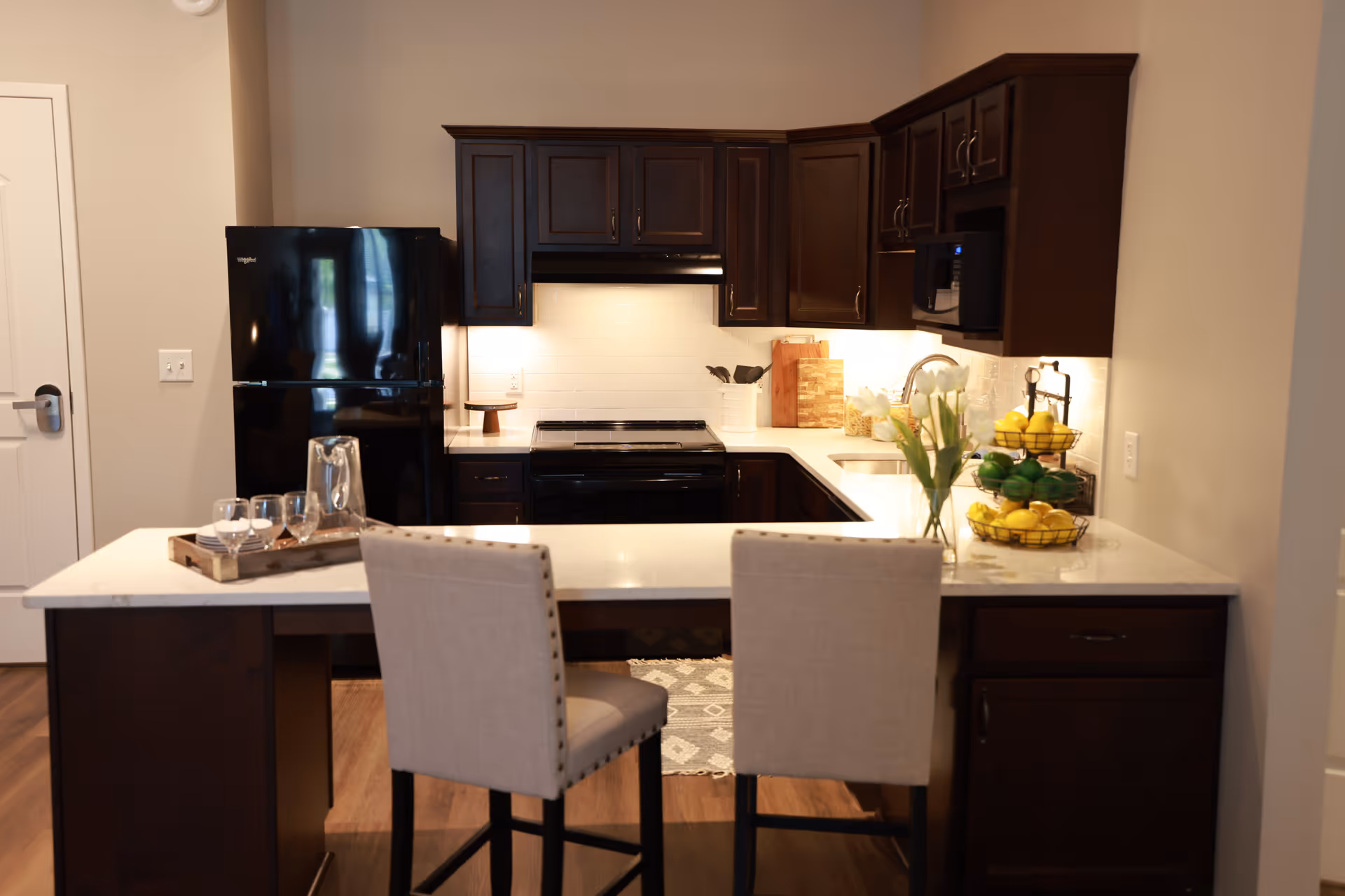 Modern kitchen with dark wood cabinets, a black refrigerator, stove, and microwave. A white countertop island with two beige upholstered chairs, a tray with glasses and a pitcher, a vase with white flowers, and a tiered fruit basket with lemons and limes are visible.