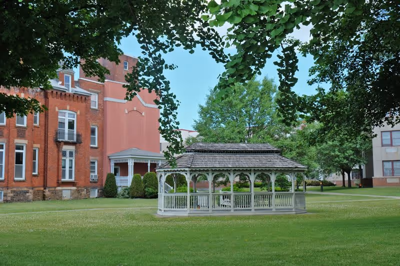 A white wooden gazebo situated on a well-maintained green lawn with trees around it. In the background, there are brick and stucco buildings with multiple windows, likely part of the Foltsbrook Center Nursing and Rehabilitation facility. The sky is clear and blue.