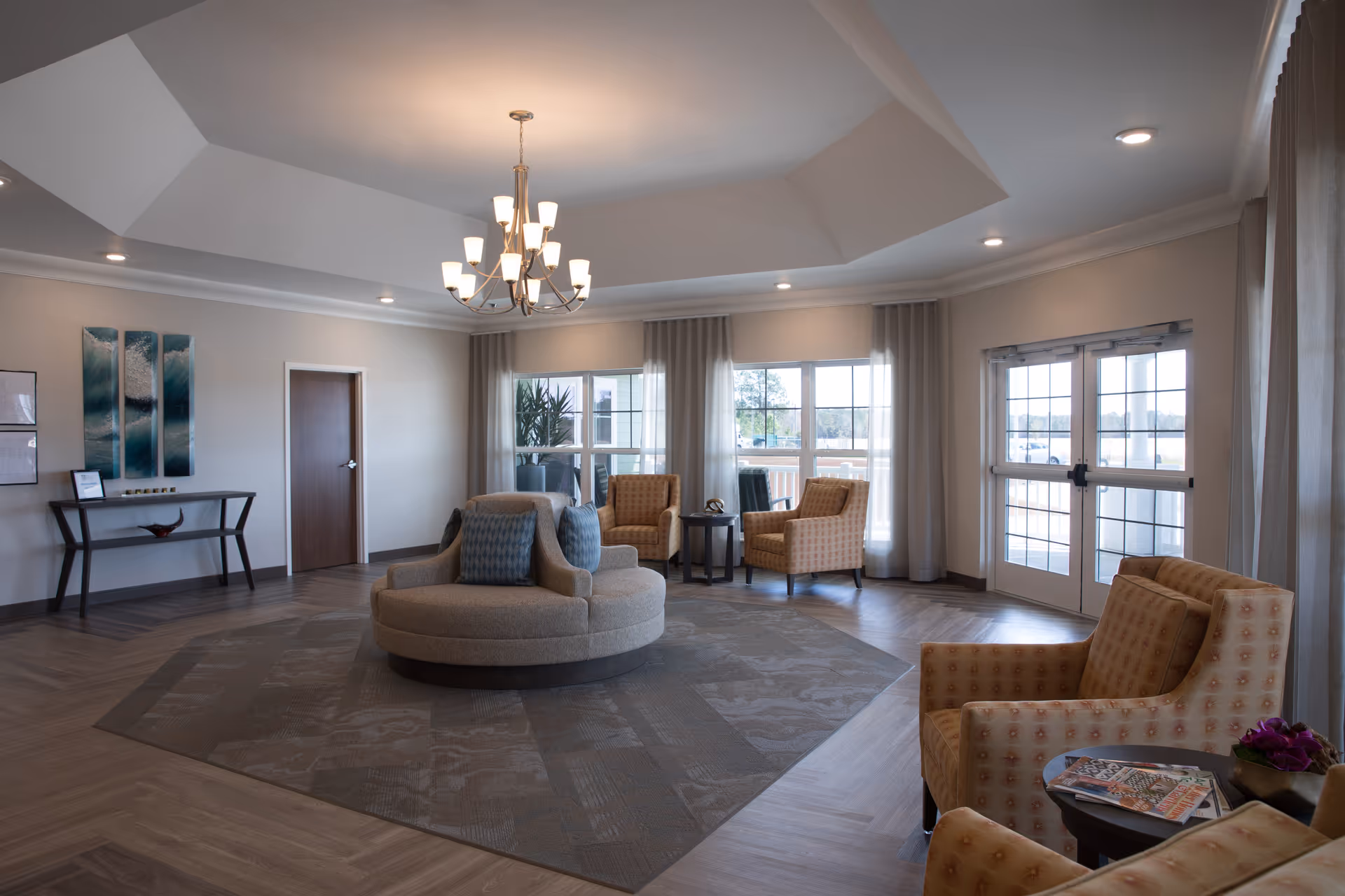 A spacious and well-lit living room area with a round beige sofa in the center adorned with blue cushions. Around the room are several patterned armchairs, a small round table with magazines and a flower pot, large windows with sheer curtains allowing natural light, a chandelier hanging from a tray ceiling, and a console table against the wall with decorative items and artwork above it.