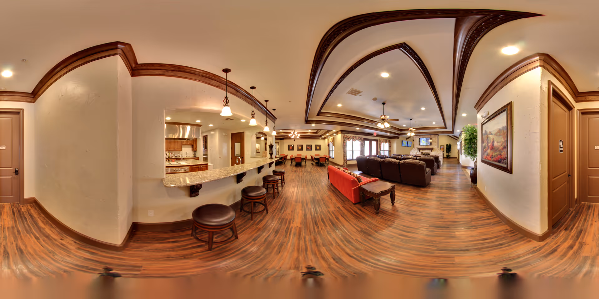 Wide panoramic view of a senior living facility common area featuring a long granite countertop with bar stools, a kitchen area with stainless steel appliances, a dining area with tables and chairs, and a living room with multiple sofas and a fireplace. The room has wooden floors, decorative ceiling beams, pendant lighting, and framed artwork on the walls.