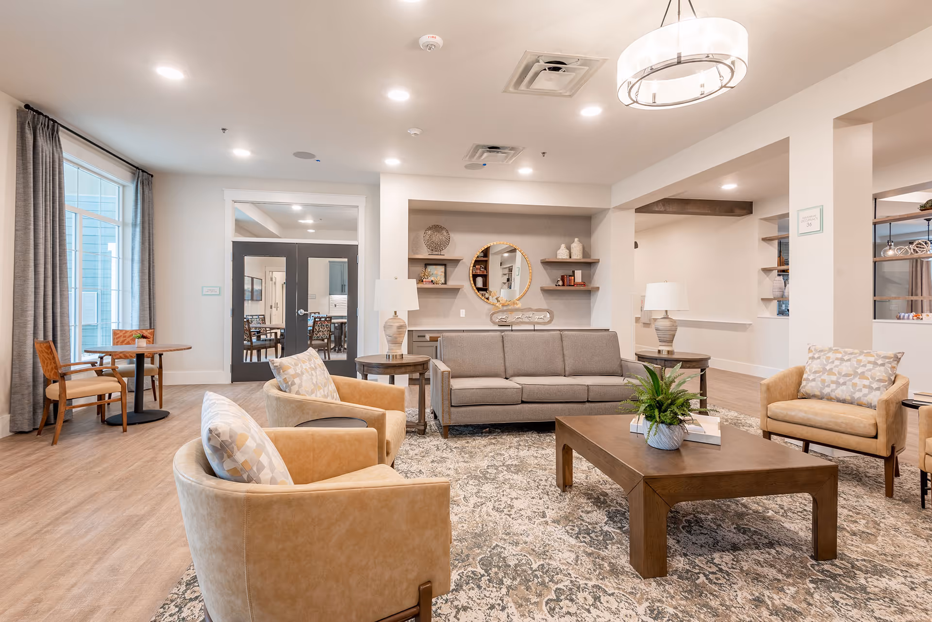A bright and spacious living room area in a senior living facility with beige armchairs and a gray sofa arranged around a wooden coffee table on a patterned rug. The room features modern lighting, a round mirror on the wall, decorative shelves with vases and books, and a small round table with chairs near a large window with gray curtains.