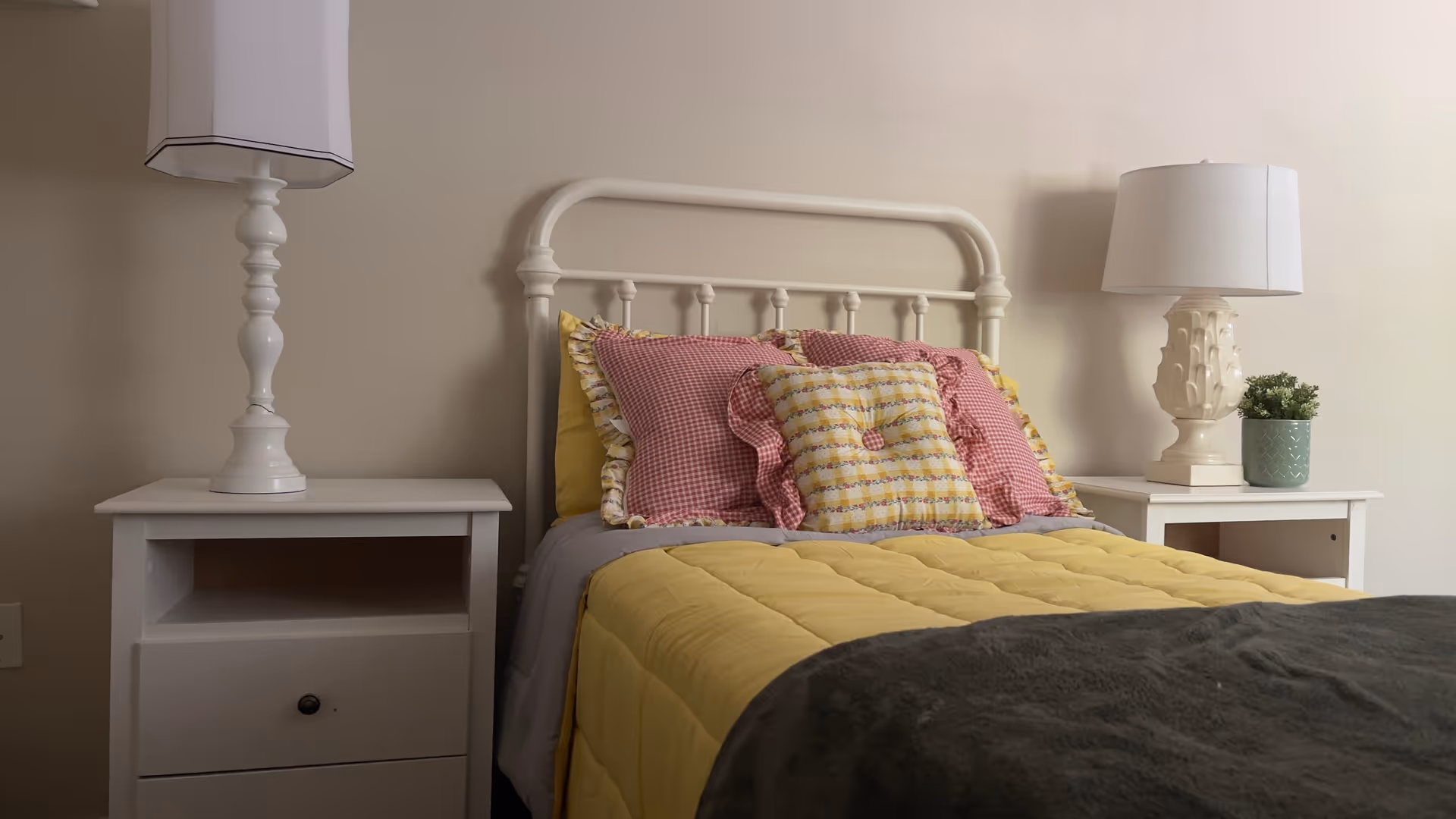 A neatly made bed with a white metal headboard, yellow quilt, and decorative pillows in yellow and red gingham patterns. There are two white nightstands on either side of the bed, each with a white lamp. A small green potted plant is on the right nightstand. The walls are beige.