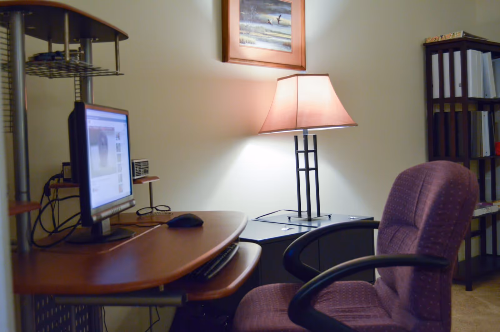 A cozy interior office corner with a computer desk and monitor, a table lamp, and an upholstered desk chair.