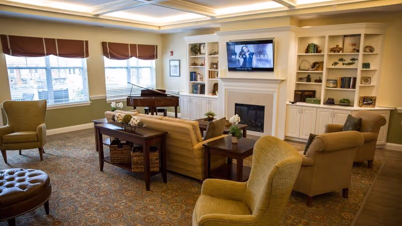 A cozy living room in a senior living facility featuring comfortable armchairs and a sofa arranged around a fireplace with a mounted flat-screen TV above it. Built-in white shelves flank the fireplace, holding books and decorative items. Large windows with brown valances allow natural light to fill the room, and a grand piano is positioned near the windows. The room has a patterned carpet and warm, neutral-colored walls.