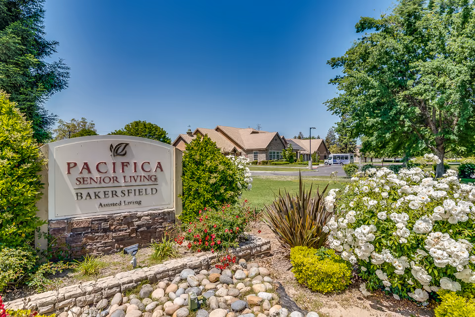 Outdoor view of Pacifica Senior Living Bakersfield assisted living facility sign surrounded by landscaping with bushes, flowers, and trees, with the facility buildings and a clear blue sky in the background.