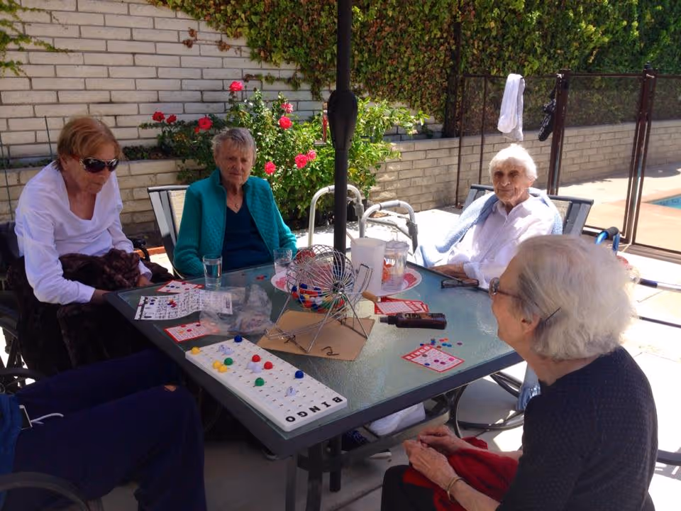 Four elderly women sitting around a glass outdoor table playing bingo. The table has bingo cards, colorful bingo chips, a bingo cage with balls, and drinks. They are seated near a pool with a safety fence, and there is a brick wall with green ivy and red flowers in the background.
