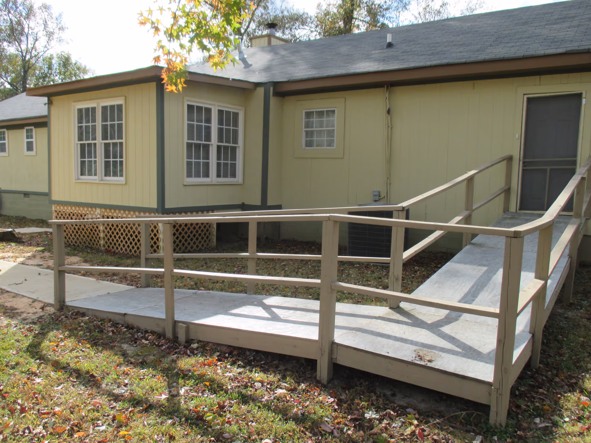 Exterior of a beige single-story building with a wooden wheelchair ramp leading to a screened door and nearby windows.