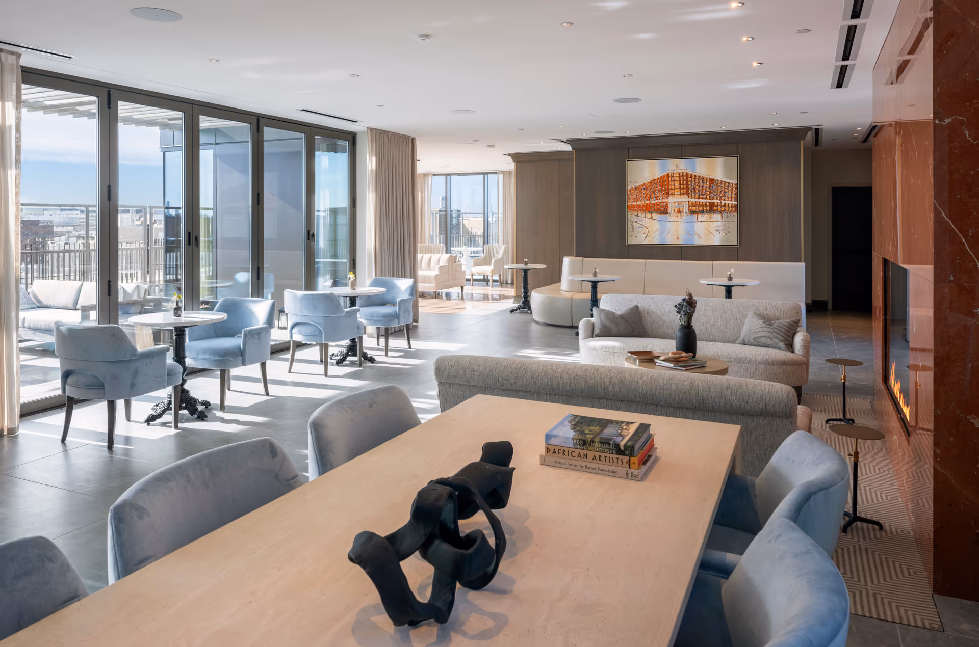 A spacious, modern living room area with large floor-to-ceiling windows letting in natural light. The room features multiple seating arrangements including light blue armchairs around small round tables near the windows, a beige sofa with cushions, and a long dining table with blue chairs. There is a fireplace with a red marble surround on the right side and a large painting on the far wall. Books and a decorative sculpture are placed on the dining table.