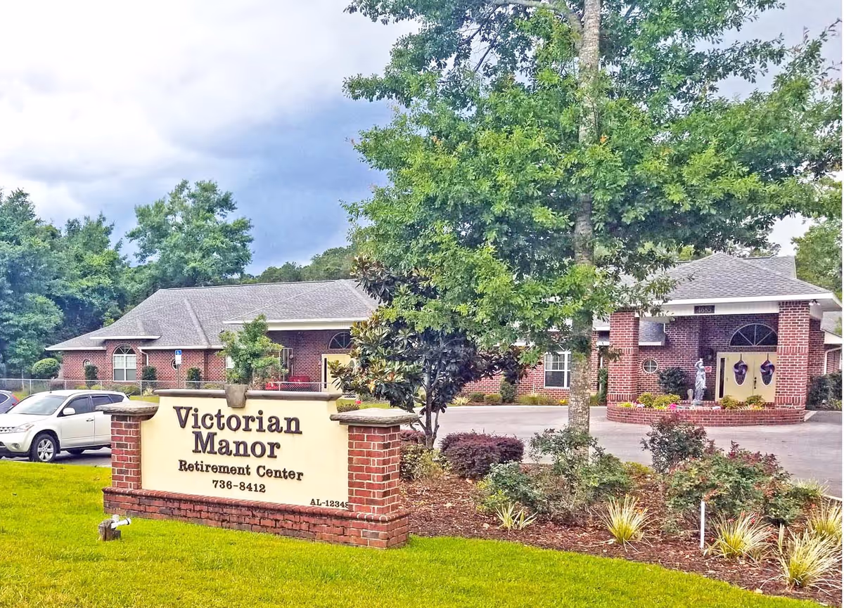 Front exterior of the Victorian Manor Retirement Center showing the brick entrance building, landscaping, parked cars, and a lawn sign reading 'Victorian Manor'.