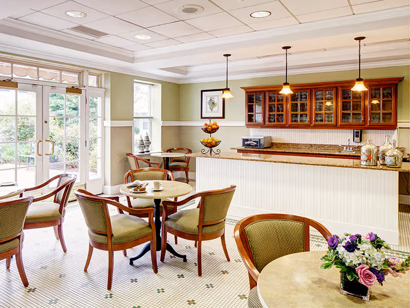 A bright and inviting common area with several round tables and wooden chairs with green upholstery. There is a counter with a granite top and wooden cabinets with glass doors above it. Three pendant lights hang from the ceiling above the counter. A tiered fruit stand and a microwave are on the counter. Large windows and glass doors let in natural light and provide a view of greenery outside. A small floral arrangement is on one of the tables in the foreground.