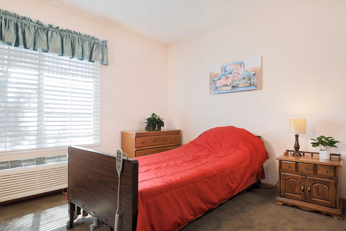 A small bedroom with a single hospital-style bed covered with a red blanket. There is a wooden nightstand with a lamp and a small plant on the right side of the bed, and a wooden dresser with another plant on top against the back wall. A window with white blinds and a green valance is on the left wall, allowing natural light to fill the room. A painting of a blue truck carrying flowers hangs on the wall above the bed.