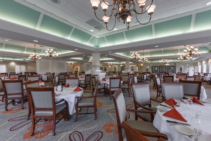 Spacious dining room with multiple round tables covered with white tablecloths, each set with plates, glasses, silverware, and red folded napkins. The room features wooden chairs with cushioned seats, a patterned carpet with floral designs, and elegant chandeliers hanging from a ceiling with recessed lighting and a greenish glow.
