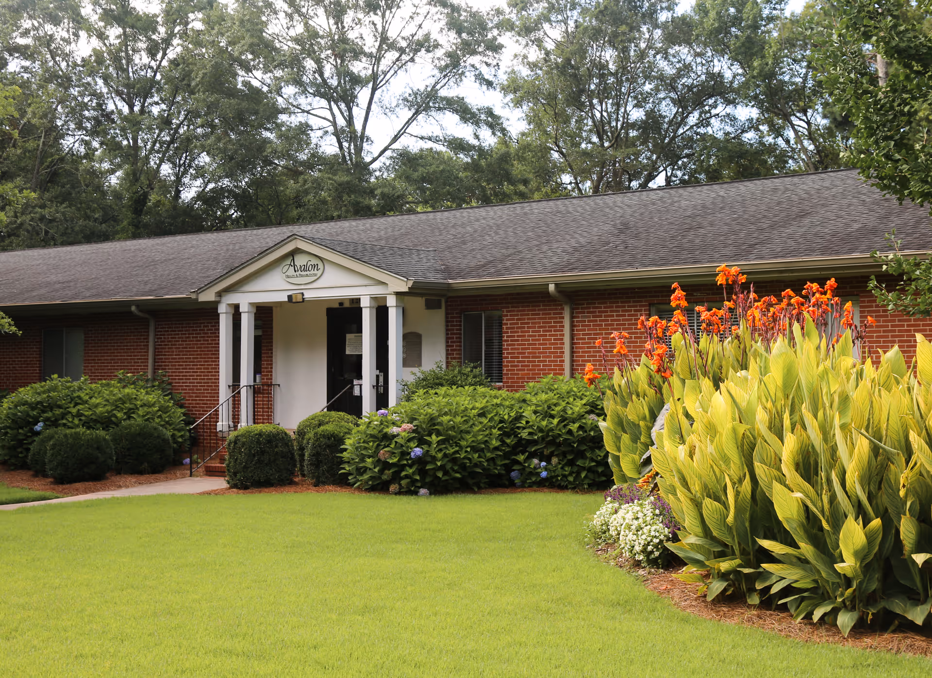 Exterior view of Avalon Health & Rehabilitation, a single-story brick building with a gabled entrance supported by white columns. The building is surrounded by well-maintained green grass, bushes, and flowering plants with tall orange blooms. Trees are visible in the background under a clear sky.