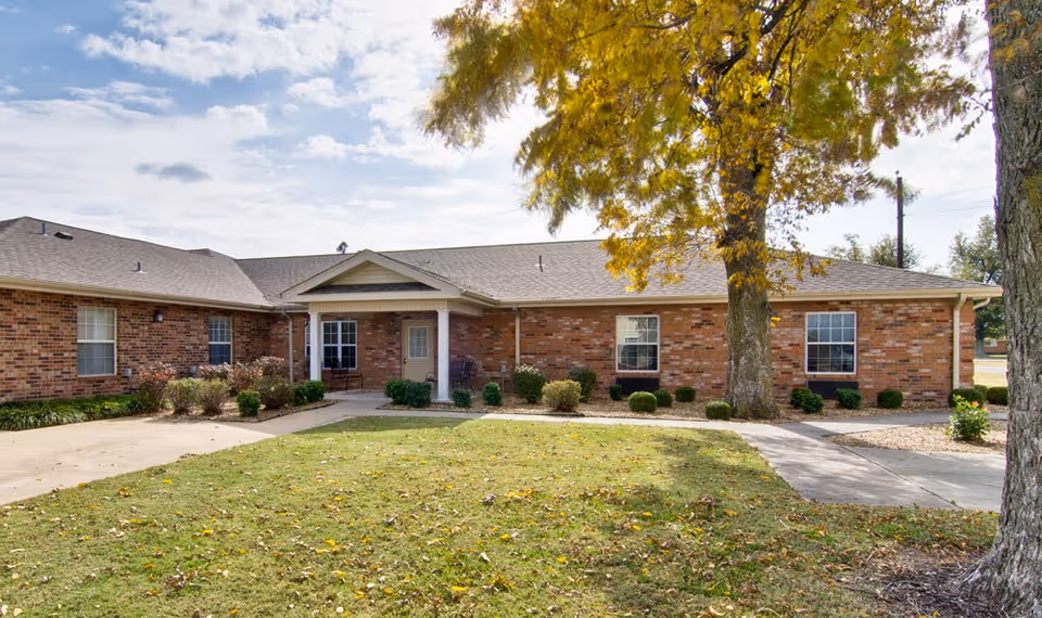 Exterior view of a single-story brick building with a covered entrance, surrounded by a lawn with scattered leaves and a large tree with yellow foliage in front.