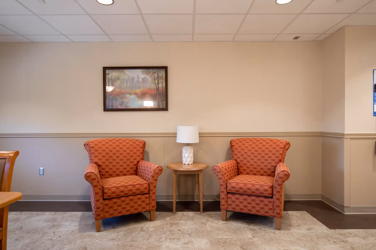 Two patterned orange armchairs with a small wooden side table and a white lamp between them, set against a beige wall with a framed landscape painting above in a well-lit room.