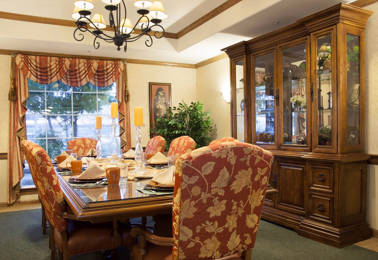 A formal dining room with a large wooden dining table set for six people. The chairs have red upholstery with a floral pattern. The table is set with plates, napkins, glasses, and three tall candles in glass holders. A large wooden china cabinet with glass doors is against the wall, displaying decorative items. A large window with patterned curtains lets in natural light, and a chandelier hangs from the ceiling.