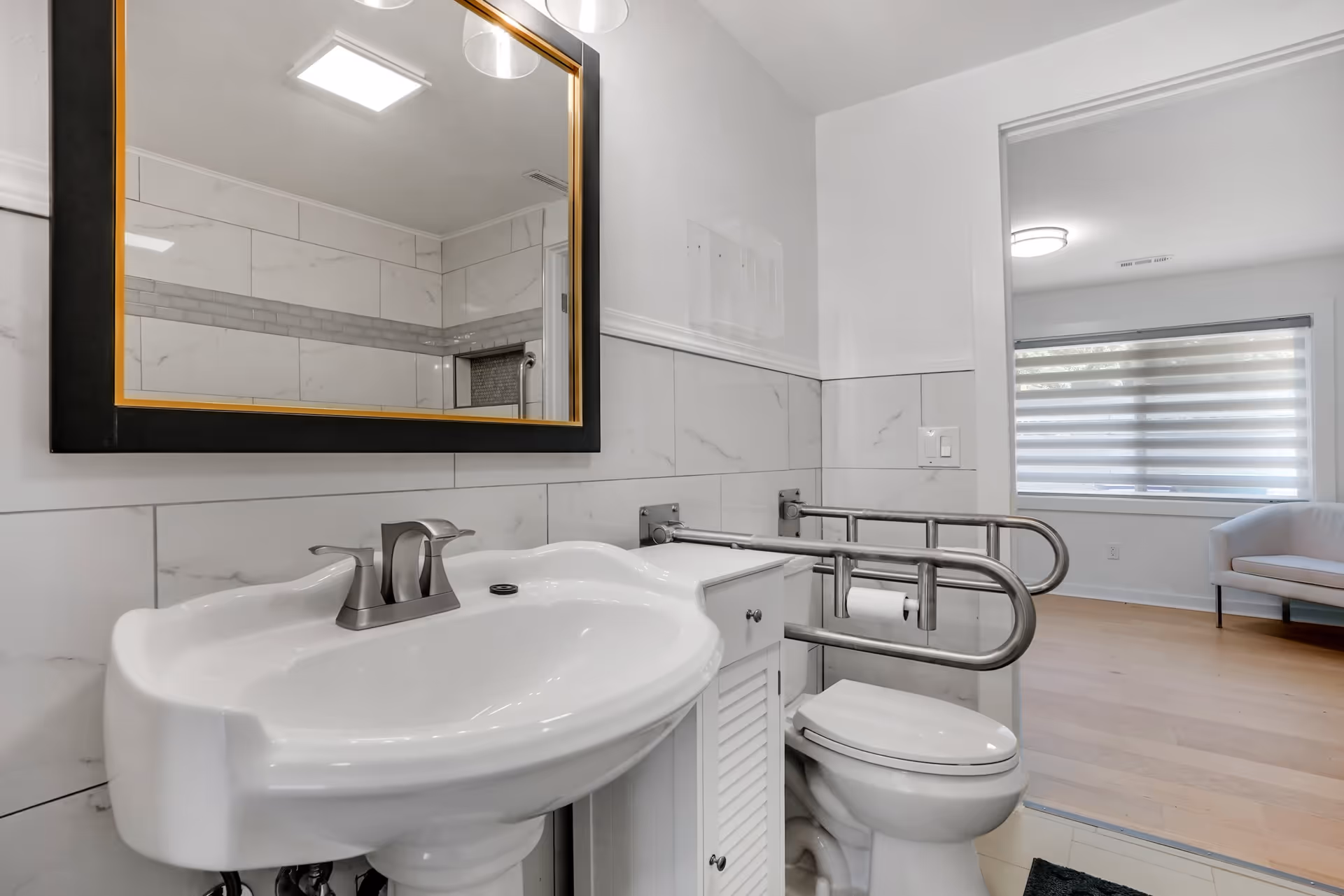 A clean and modern bathroom with a white pedestal sink, a large mirror with a black and gold frame, a toilet with stainless steel grab bars, and white tiled walls. The bathroom opens into a room with wooden flooring and a white armchair near a window with horizontal blinds.
